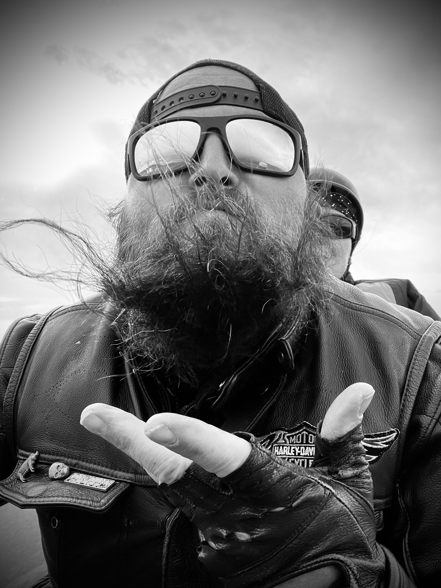 A black and white photograph of a man with a long beard, wearing sunglasses and a Harley-Davidson leather vest. He is hold...